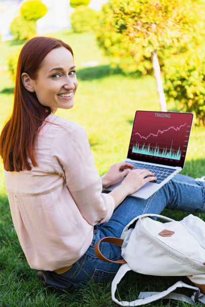 smiling attractive woman using laptop with traiding appliance in park and looking at camera - Photo, Image