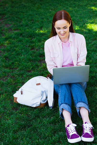 attractive freelancer using laptop and sitting on green grass in park - Фото, зображення