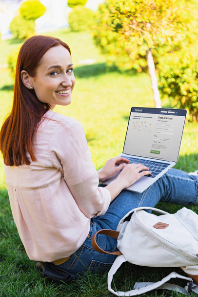 smiling attractive woman using laptop with loaded facebook page in park and looking at camera - Foto, immagini