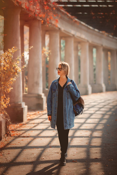 Mulher elegante em jaqueta de ganga e óculos andando no beco com colunas. Hora da época de outono
.  - Foto, Imagem