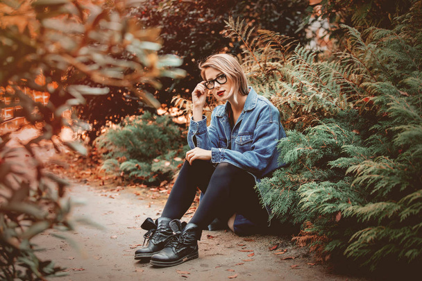 Giovane donna in giacca di jeans e occhiali seduta a terra nel parco. Scena della stagione autunnale. Tendenze hipster stile vestiti
. - Foto, immagini