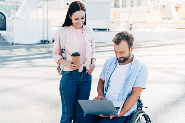 smiling handsome boyfriend in wheelchair using laptop and girlfriend standing with coffee in paper cup on street - Фото, зображення
