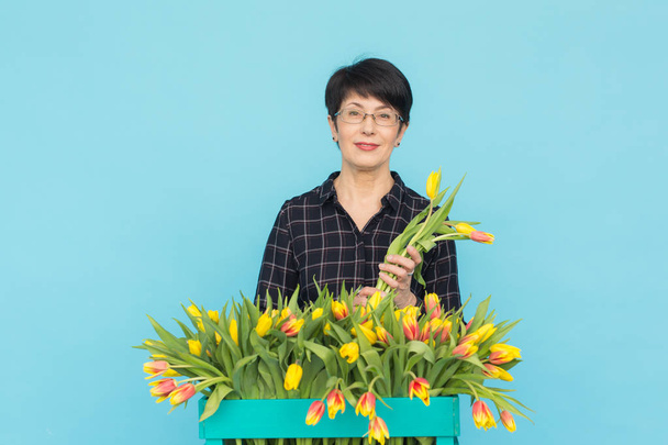 glückliche Frau mittleren Alters mit Brille, die eine Schachtel Tulpen vor blauem Hintergrund hält - Foto, Bild