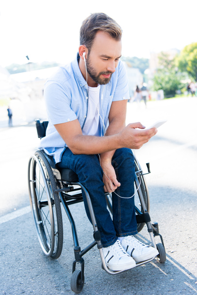 handsome man in wheelchair listening to music with smartphone on street - Photo, Image