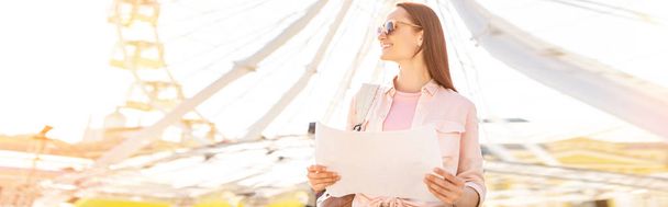 panoramic view of attractive tourist in sunglasses standing with map near observation wheel - Photo, Image