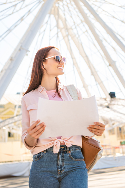 smiling attractive tourist in sunglasses standing with map near ferris wheel in amusement park and looking away - Photo, Image