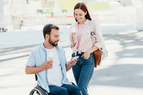 handsome boyfriend in wheelchair and girlfriend with ice cream using smartphone on street - Photo, Image