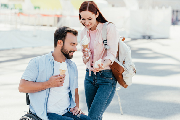 handsome boyfriend in wheelchair and girlfriend with ice cream looking at smartphone on street - Foto, afbeelding