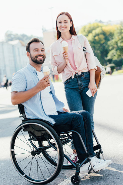 χαμογελαστός, όμορφος ο φίλος σε αναπηρικό καροτσάκι και φίλη με παγωτό βλέπουν φωτογραφική μηχανή στην οδό - Φωτογραφία, εικόνα