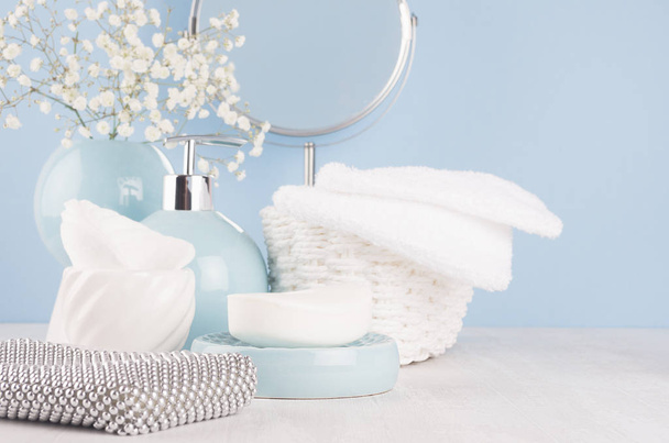 Products for body and skin care in light blue ceramic bowls, silver cosmetic bag, circle mirror and white flowers on wood table and pastel blue wall, closeup. - Foto, Imagen