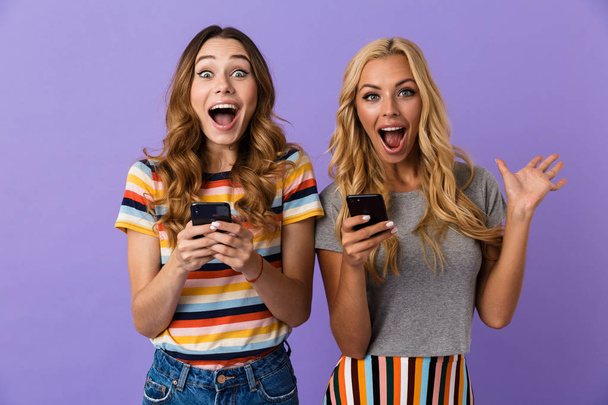 Two pretty excited young girls friends standing isolated over violet background, using mobile phones - Foto, Imagem