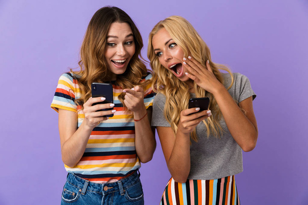 Two pretty cheerful young girls friends standing isolated over violet background, using mobile phones - Фото, зображення