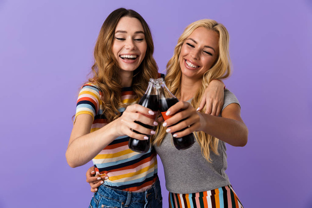 Two pretty cheerful young girls friends standing isolated over violet background, showing glass bottles with fizzy drink, toasting - Foto, immagini