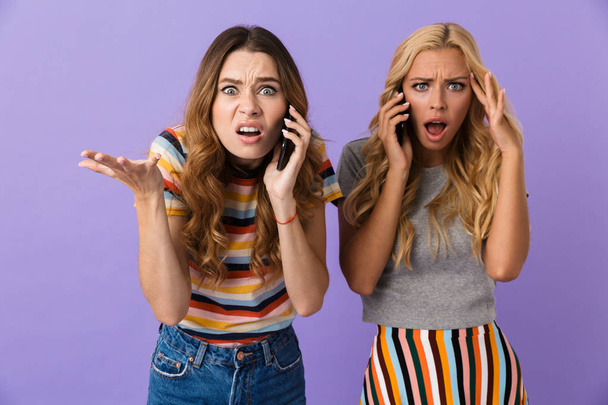Two pretty confused young girls friends standing isolated over violet background, talking on mobile phones - Фото, зображення