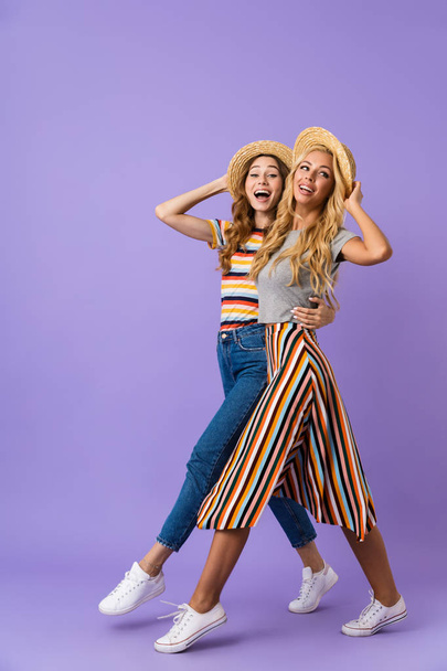 Full length of two pretty young girls friends in summer hats walking isolated over violet background, having fun - Foto, Imagem