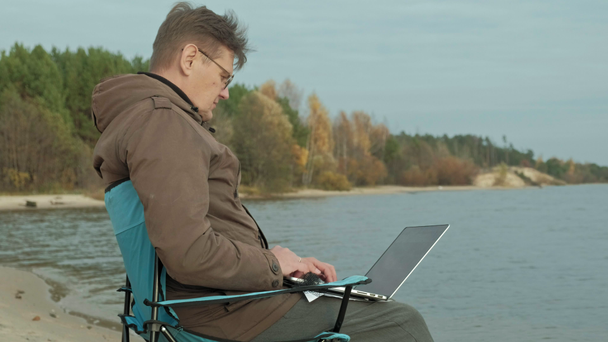 Uomo d'affari maturo seduto e rilassante vicino a un fiume. Uomo in una giacca calda e utilizzando il computer portatile
. - Filmati, video