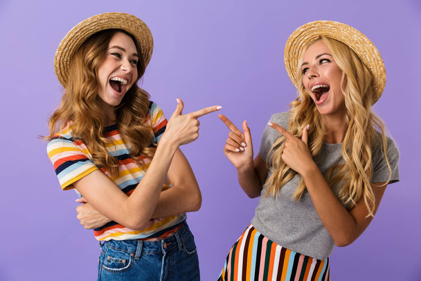 Two pretty young girls friends in summer hats standing isolated over violet background, pointing fingers at each other - Photo, Image