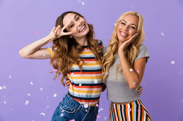 Two cheerful young girls friends standing isolated over violet background, showing peace under confetti shower - Foto, immagini