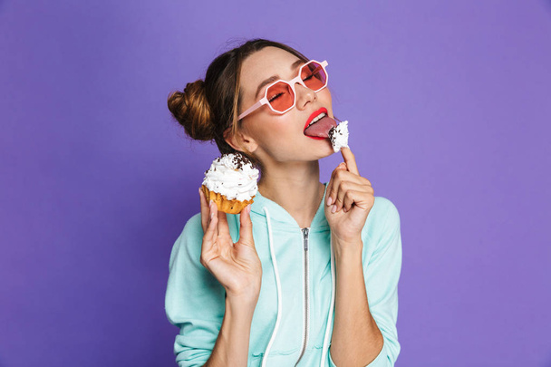 Portrait of a seductive young girl with bright makeup over violet background, eating cupcake - Fotó, kép