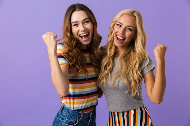 Two pretty young girls friends standing isolated over violet background, celebrating success - Foto, Bild