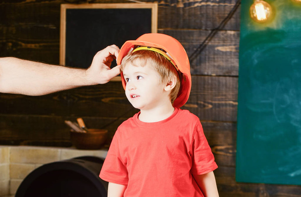 Toddler in protective hard hat, helmet at home in workshop. Child cute and adorable stands while male hand putting hard hat on head. Protection and safety concept. Carefully protect kid with helmet - Fotoğraf, Görsel