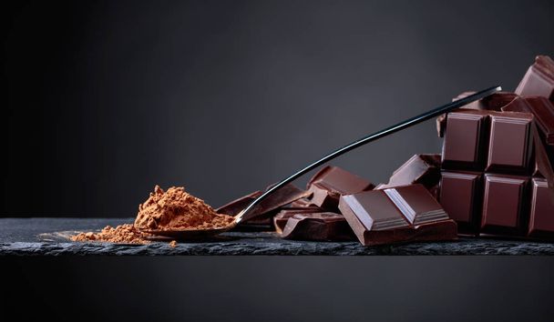 Gebroken chocoladestukjes en poeder van de cacao in kleine lepel op een zwarte achtergrond. Ruimte voor uw tekst kopiëren. - Foto, afbeelding