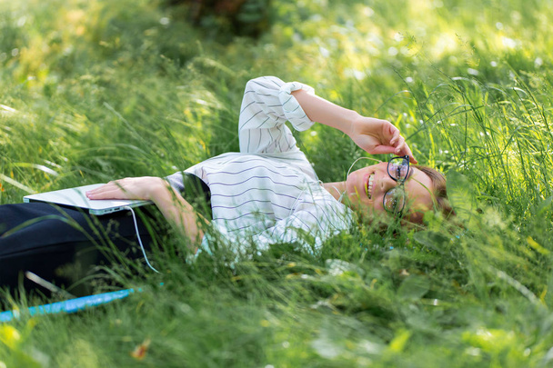 Nainen lepää puistossa nurmikolla kannettavan tietokoneen kanssa ja kuuntelee musiikkia.
 - Valokuva, kuva