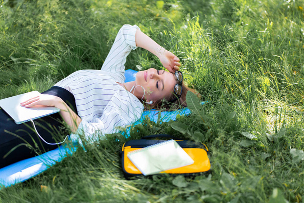 Woman resting in the park lying on the grass with a laptop and listening to music - Photo, Image
