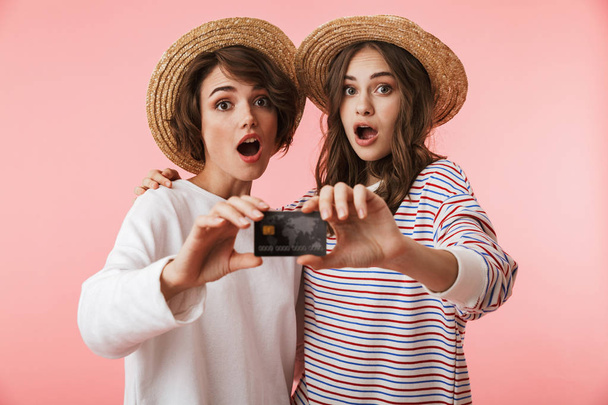 Image of excited young women friends isolated over pink wall background holding credit card. - 写真・画像