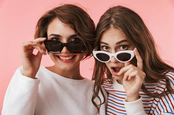 Image of shocked emotional young women friends isolated over pink wall background wearing sunglasses. - Foto, immagini