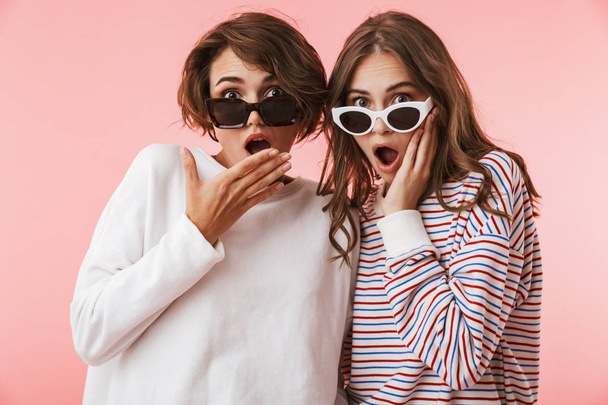 Image of shocked emotional young women friends isolated over pink wall background wearing sunglasses. - Fotó, kép