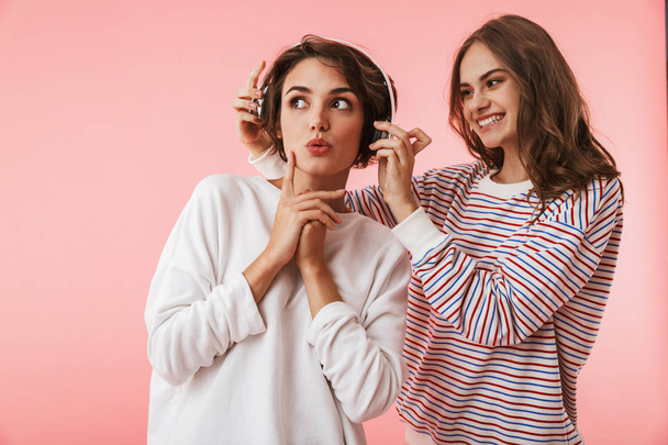 Image of happy emotional young women friends posing isolated over pink background listening music with headphones. - Foto, immagini