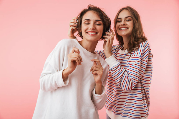 Image of happy emotional young women friends posing isolated over pink background listening music with headphones. - Фото, изображение