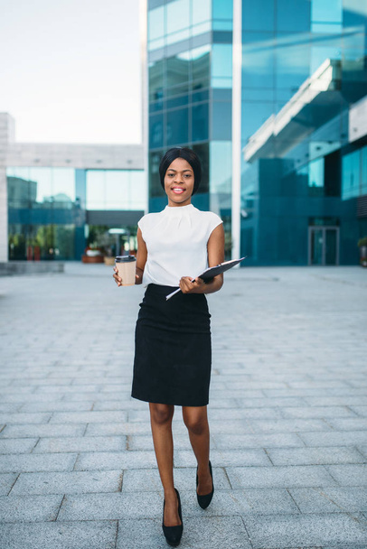 Geschäftsfrau mit Kaffeetasse und Notizblock aus Pappe im Freien, Bürogebäude im Hintergrund. schwarze Geschäftsfrau in Rock und weißer Bluse - Foto, Bild