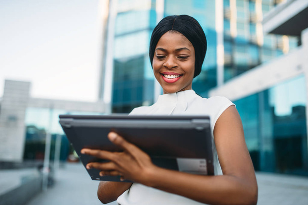 İş kadını dizüstü bilgisayar ekranında ofis binası önünde görünüyor. Beyaz bluz, siyah iş kadını gülümseyen pc açık havada üzerinde çalışır - Fotoğraf, Görsel