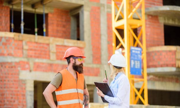 Discuss progress project. Safety inspector concept. Woman inspector and bearded brutal builder discuss construction progress. Construction project inspecting. Construction site safety inspection - 写真・画像