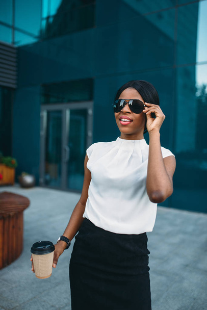 Cheerful business woman in sunglasses with cardboard coffee cup resting during the break outdoors, office building on background. Black businesswoman in skirt and white blouse - Zdjęcie, obraz