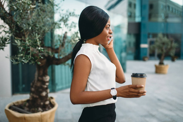 Jonge zakenvrouw met kartonnen koffiekopje en mobiele telefoon tegen kantoorgebouw op achtergrond. Glimlachend zwarte zakenvrouw in rok en witte blouse - Foto, afbeelding