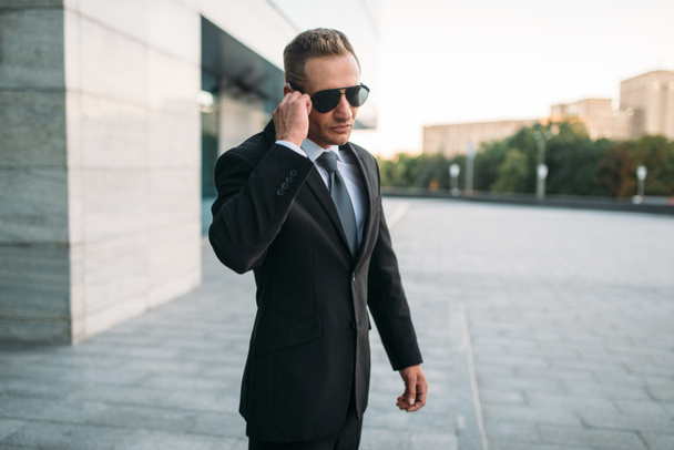 Αρσενικό σωματοφύλακας στο κοστούμι και τα γυαλιά ηλίου που μιλάει με ασφάλεια το ακουστικό σε εξωτερικούς χώρους. Επαγγελματική φύλαξη είναι ένα επικίνδυνο επάγγελμα - Φωτογραφία, εικόνα