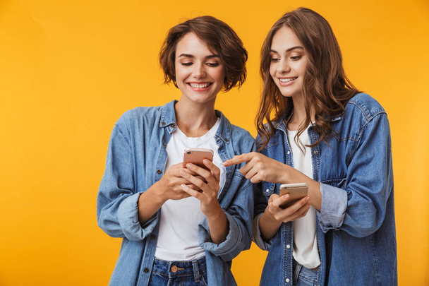 Photo of cheerful happy emotional young women friends posing isolated over yellow background using mobile phones. - Fotoğraf, Görsel