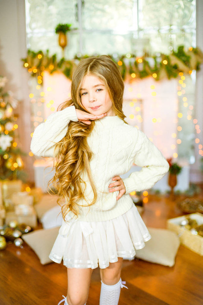 Cute little girl with blond long curly hair in a light knitted sweater by the festive fireplace with garlands and candles - Fotó, kép