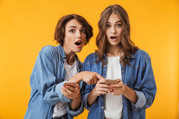 Photo of shocked emotional young women friends posing isolated over yellow background using mobile phones. - Foto, imagen