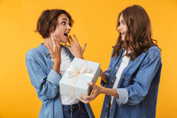 Image of cheerful happy women friends isolated over yellow wall background holding surprise gift box present. - Foto, Imagem