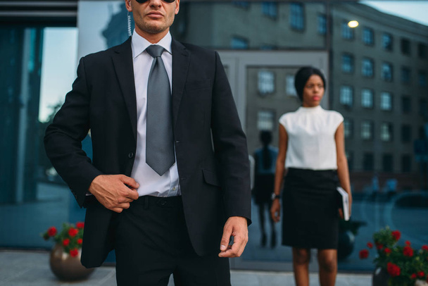 Bodyguard in suit and sunglasses, female VIP client on background. Security guard is a risky profession, professional guarding, business persons protection - Photo, Image