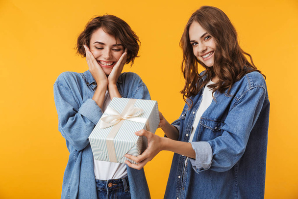Image of cheerful happy women friends isolated over yellow wall background holding surprise gift box present. - Foto, imagen