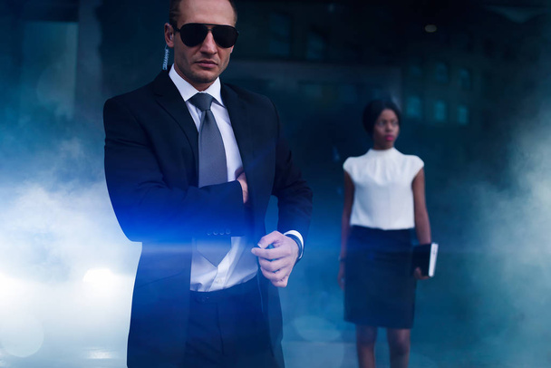 Serious bodyguard in suit, sunglasses and earpiece, female client on background. Security guard is a risky profession, professional guarding of business persons - Фото, изображение