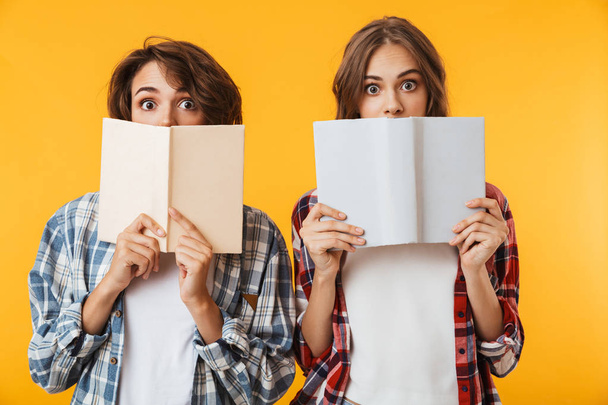 Photo of emotional young women friends posing isolated over yellow background holding books covering faces. - Fotoğraf, Görsel