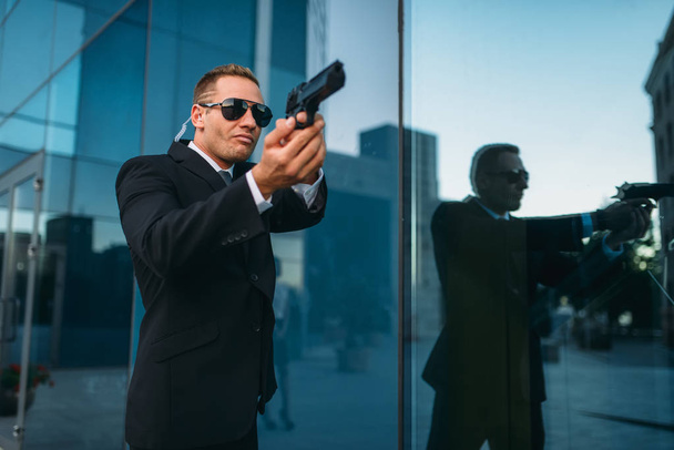 Bodyguard in suit and sunglasses with security earpiece and gun in his hands. Guarding is a risky profession, politicians and business persons protection from the danger of life - Photo, Image