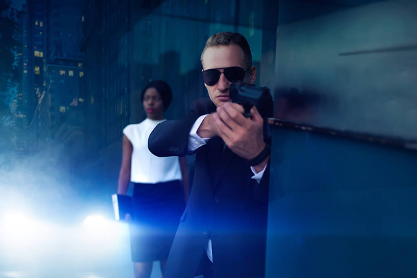 Bodyguard in suit and sunglasses with a gun in his hand protect female VIP client. Security guard is a risky profession, professional guarding, business persons protection from the danger of life - Photo, Image