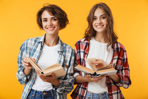 Photo of young women friends posing isolated over yellow background holding books. - Foto, afbeelding